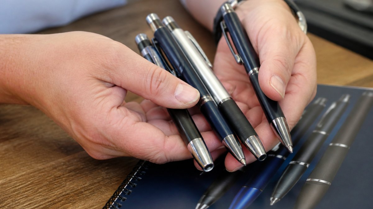 Hands holding high-quality promotional pens with metal trims and rubberized grips resting on a notebook.