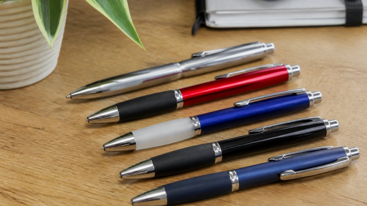 Close-up of several branded promotional pens displayed on a desk, showing different finishes including metallic, frosted barrels, rubber grips, and chrome trim details with custom logos visible